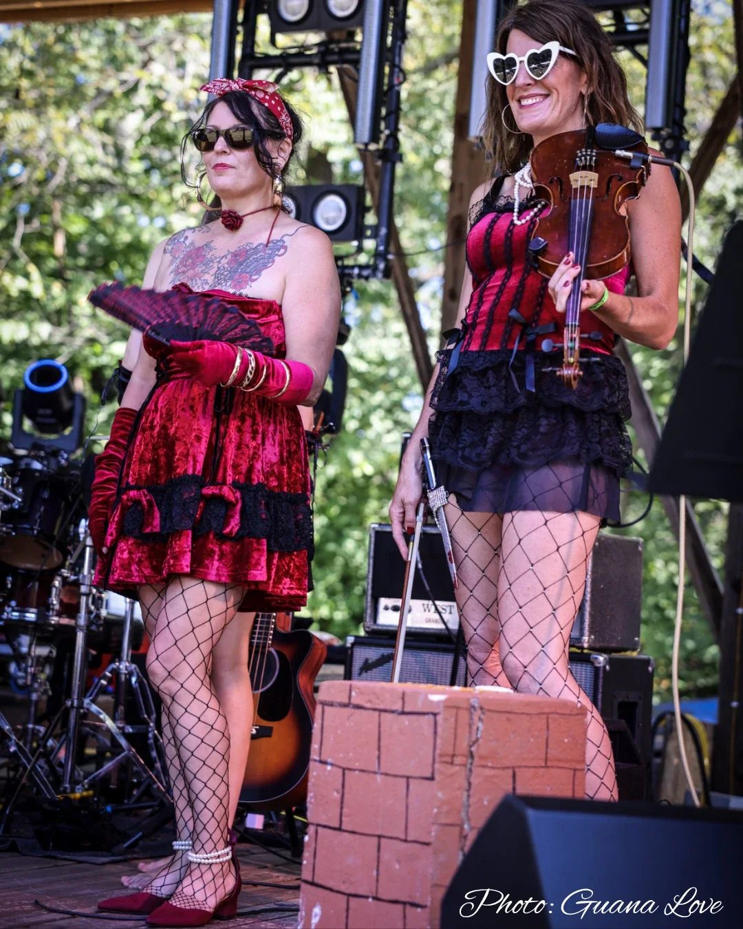 Jenny holding a fan beside the fiddle player in heart-shaped sunglasses on an outdoor stage